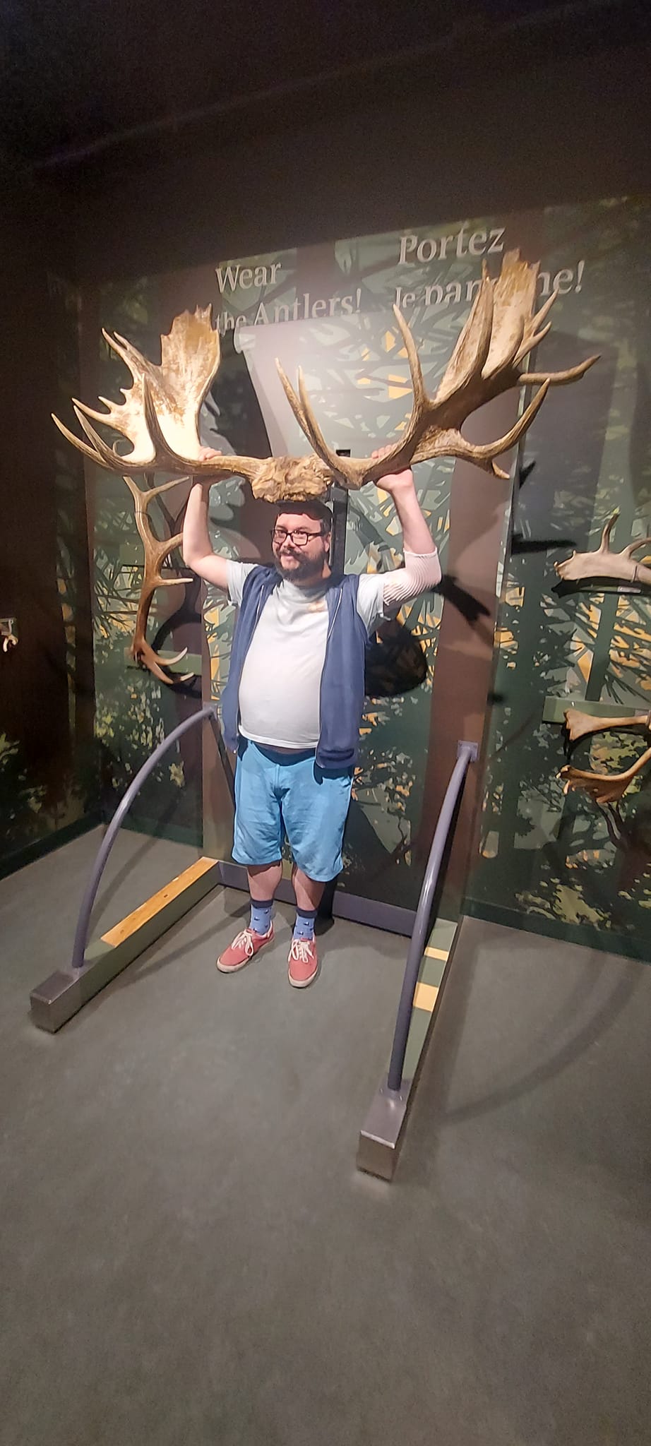 A man with a ball cap and bushy facial hair is standing with a large set of moose antlers on his head, they are as wide as he is tall